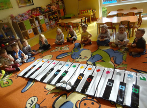 Dzieci siedzą wokół rozłożonego na dywanie instrumentu Kolorpiano.